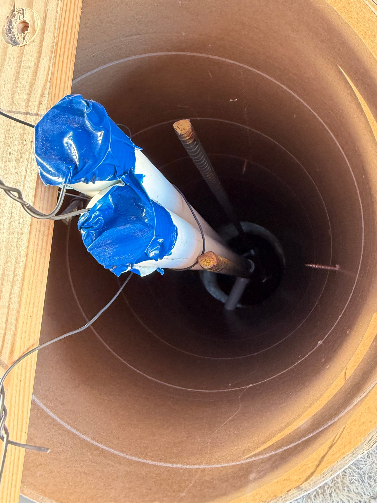 Looking down into a cardboard form tube at rebar and two white conduits capped with blue tape, hanging on tie wire.