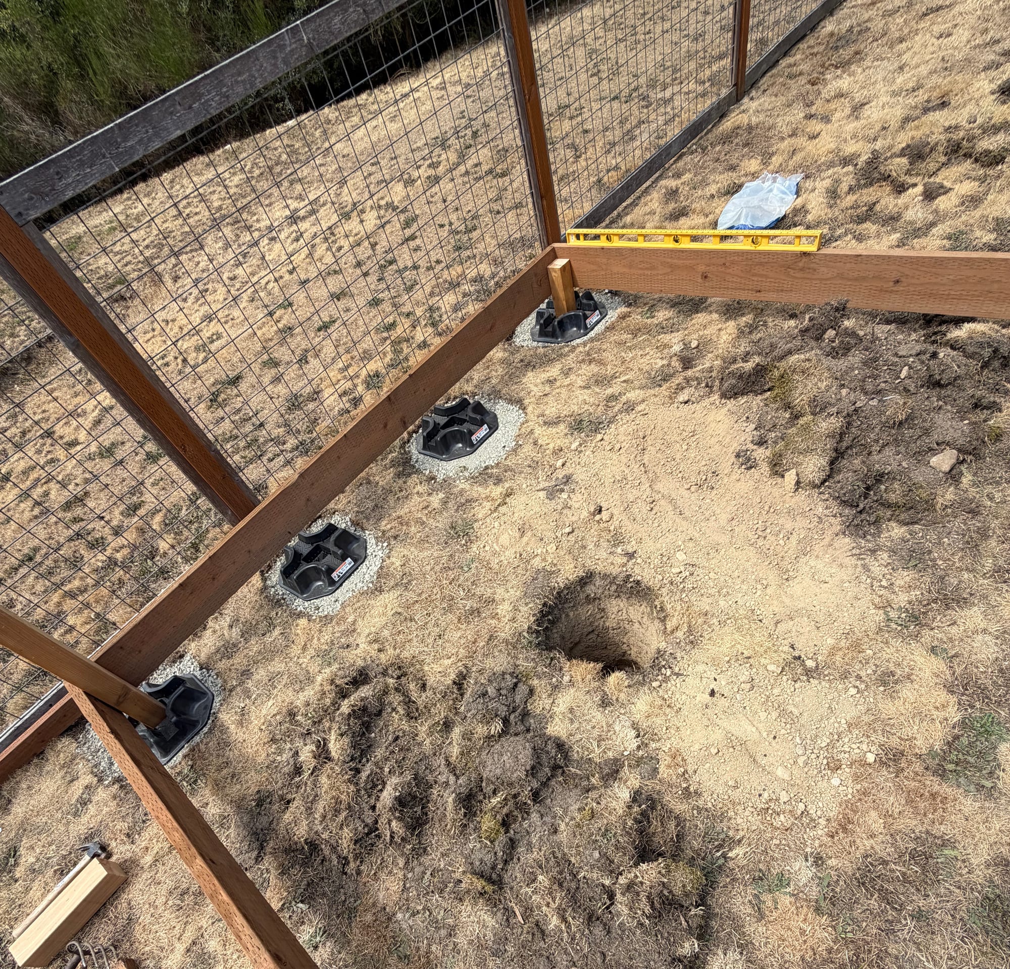 Black plastic deck blocks on gravel pads in a line along a fence, a yellow level on a board across them, and a round hole dug in the soil.