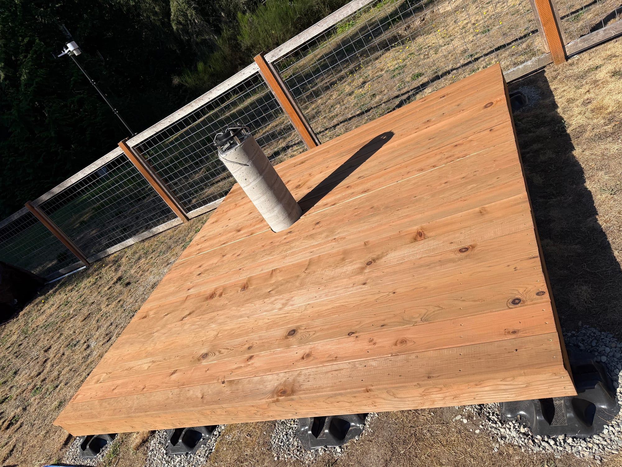 A square wooden deck of long boards on black plastic blocks, with a concrete pier rising through an opening near one side, next to a wire fence.