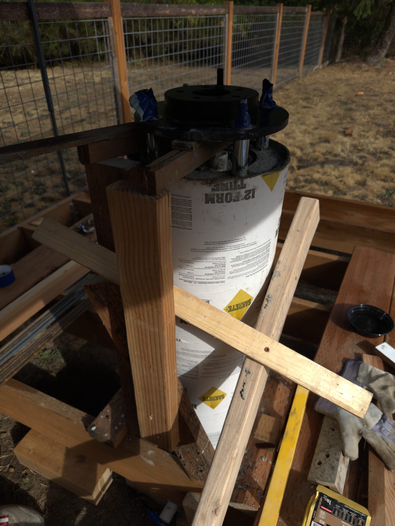 A timber frame of uprights and diagonal braces around a concrete-filled form tube, holding the black brake-rotor adapter on the fresh concrete.