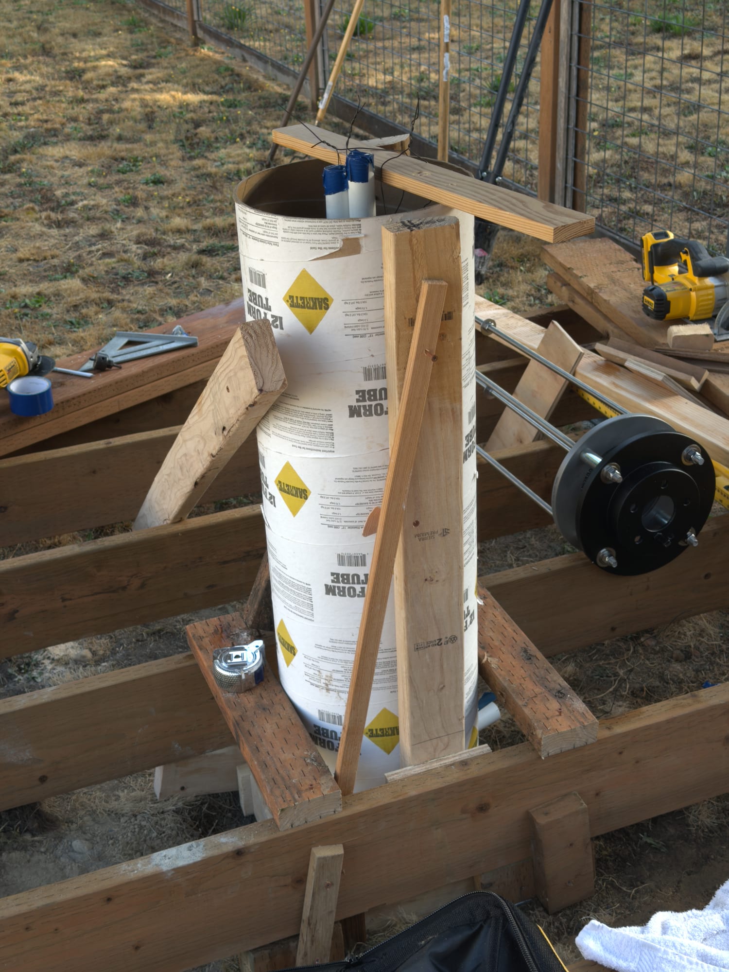 A cardboard concrete form tube braced with timber inside deck framing, two taped conduit ends at its rim under a board, and the brake-rotor adapter lying on the framing beside it.