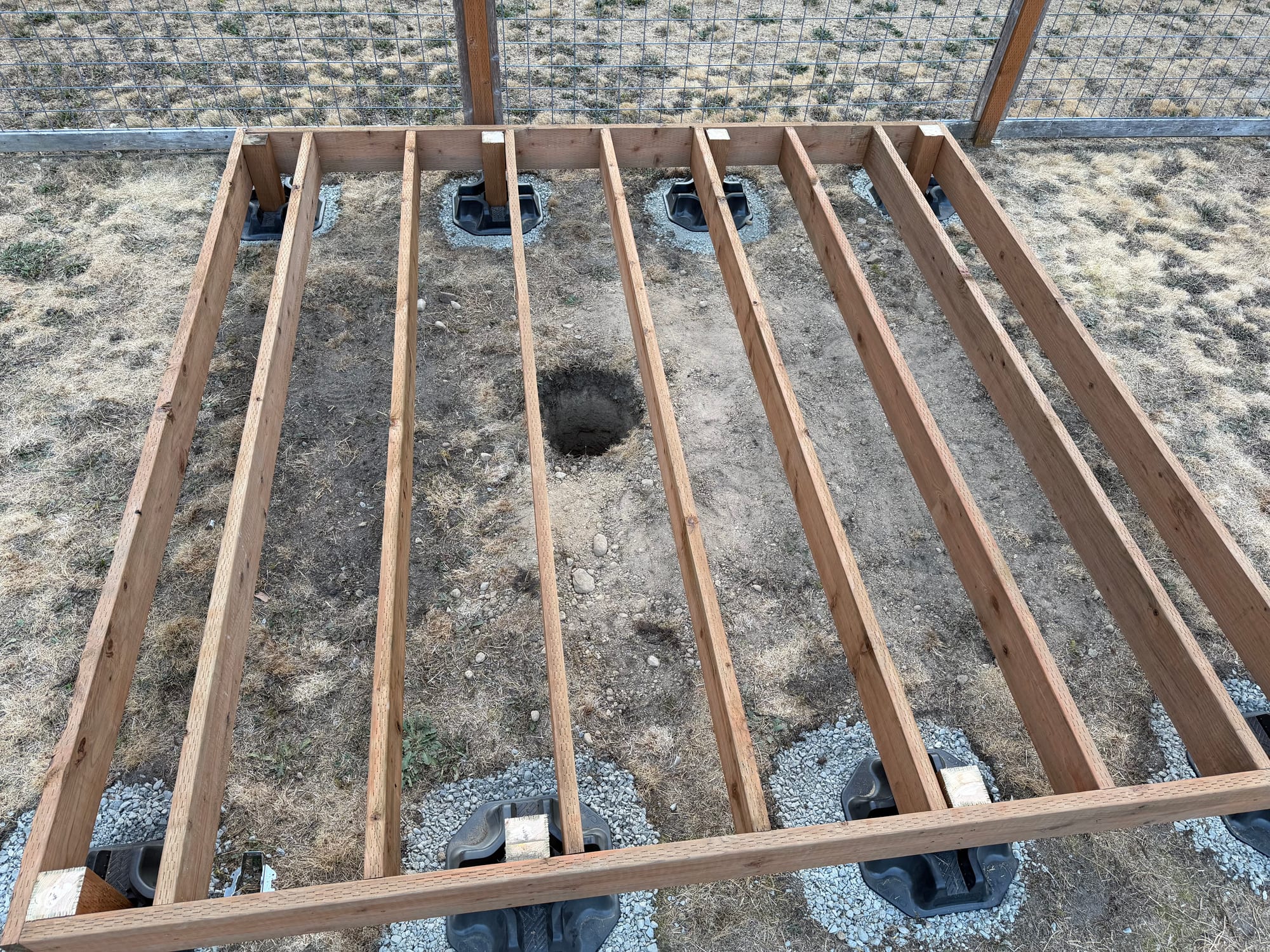 Parallel floor joists inside a rectangular perimeter frame seen from above, with a round hole dug in the soil between two of them and deck blocks under the edges.
