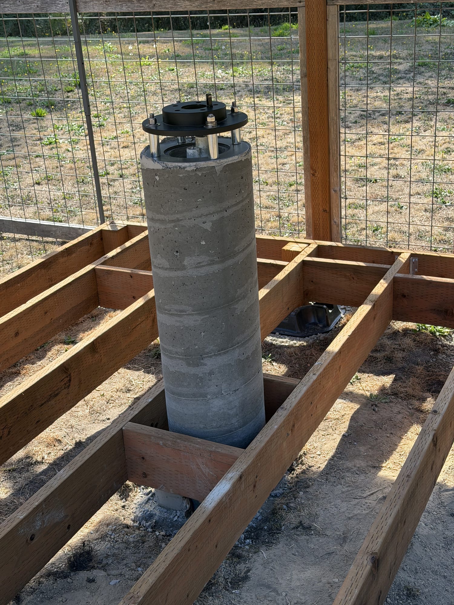 A concrete pier with the cardboard form removed and the brake-rotor adapter on top, standing in an opening in the deck framing.