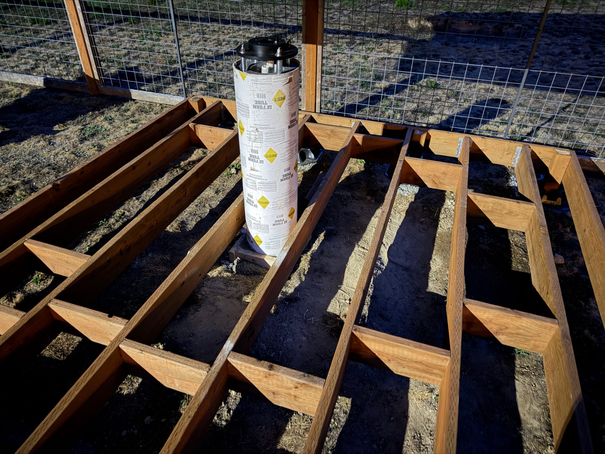 Deck framing with short blocking pieces between the joists at staggered positions, and a cardboard concrete form standing in the pier opening.