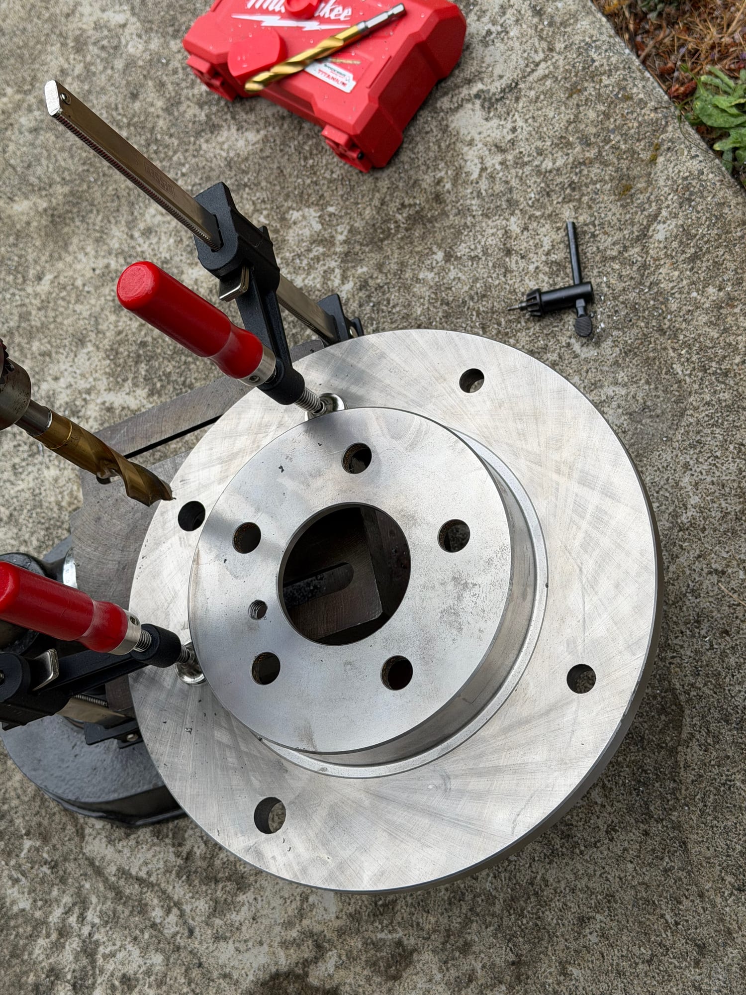 A machined brake rotor clamped with two bar clamps to a drill-press table, with a stepped drill bit in the chuck.
