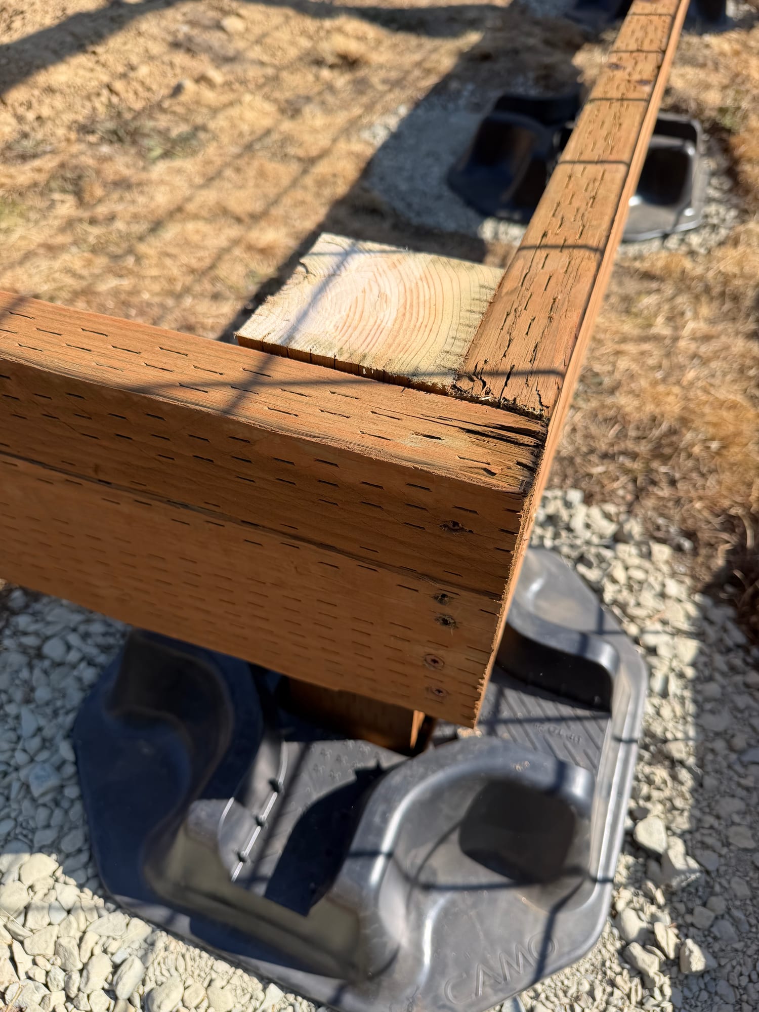 A corner where two pressure-treated boards meet, above a black plastic CAMO deck block set in crushed gravel.