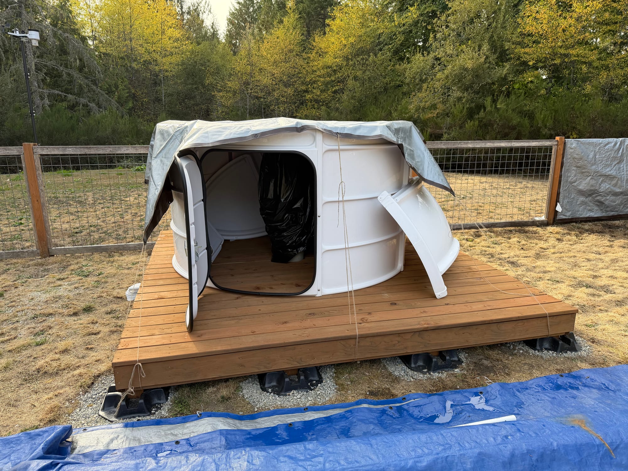 A ring of curved white wall panels with its door open, standing on a wooden deck under a gray tarp, with a curved white panel leaning against it.